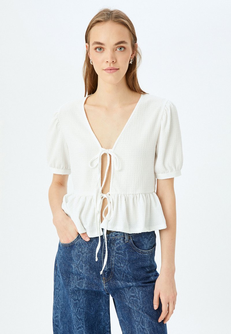 Blusa bianca texturizzata con maniche corte a palloncino, scollatura a V e chiusura con lacci sul davanti. L'orlo a peplum aggiunge volume e forma al design.