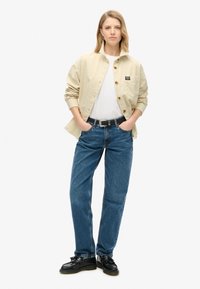 Chemise beige à boutons avec une poche, associée à un jean droit bleu et des chaussures noires. La tenue présente un design simple et décontracté.