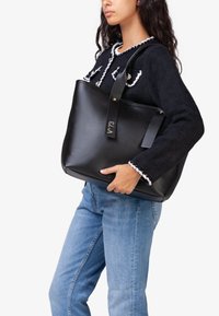 Zwarte leren totebag met een gestructureerde vorm, dubbele handvatten bovenaan en een gouden geïmpregneerd label. Heeft een gladde textuur en een minimalistisch ontwerp.