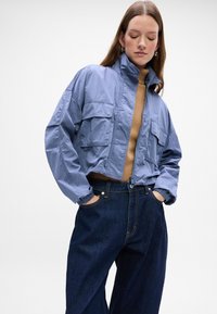 Chaqueta corta azul con cuello alto, dos bolsillos en el pecho y puños elásticos, usada sobre una blusa color beige y pantalones de mezclilla oscuros de pierna ancha.