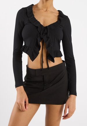 Femme porte un top court noir à manches longues avec des volants et un lien à nouer à l'avant, associé à une mini-jupe asymétrique noire.