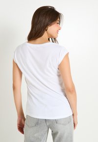 Camiseta de algodón blanca de manga corta y cuello redondo, vista por detrás. Combinada con unos jeans de mezclilla gris claro.