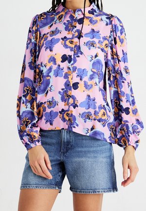 Femme portant un chemisier rose avec un imprimé floral violet et orange, manches longues bouffantes, et un short en jean.
