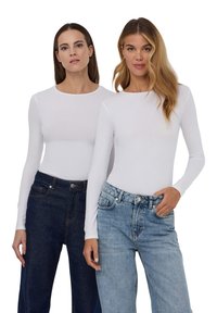 Due donne indossano top bianchi a manica lunga. Una ha jeans in denim scuro, mentre l'altra ha jeans chiari. Entrambi i top hanno un collo arrotondato.
