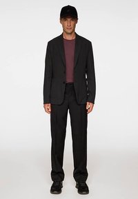 Svart skräddarsydd kavaj över en vinröd skjorta, kombinerad med svarta byxor och svarta sneakers. En svart keps kompletterar outfiten. Slätt tyg, elegant design.