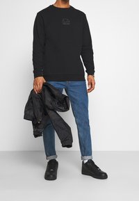 Hombre vestido con sudadera negra y jeans azules arremangados sosteniendo una chaqueta negra, de pie frente a un fondo blanco liso.