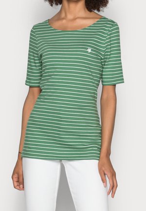 Vrouw draagt een groen-wit gestreept shirt met korte mouwen en witte broek, staande tegen een effen achtergrond.