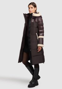 Parka lunga di colore marrone scuro con cappuccio foderato in pelliccia, tasche con zip e polsini a costine. La texture è liscia e imbottita.