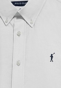 Vit skjorta med krage i ett texturerat tyg, med klara knappar och en liten marinblå broderad figur på vänster bröst.