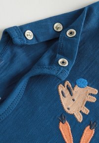 Chemise en coton bleu ornée d'un personnage brodé portant un chapeau rond. Fermetures à pression au col et détail en tissu texturé visibles.