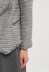 Blazer de cuadros en blanco y negro con textura, bordes deshilachados y un bolsillo con cremallera. Llevado sobre una tela gris por debajo, mostrando un estilo en capas.