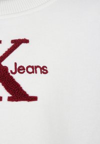 Sudadera blanca con una gran letra "K" texturizada en burdeos y la palabra "Jeans" bordada en una fuente suave debajo.