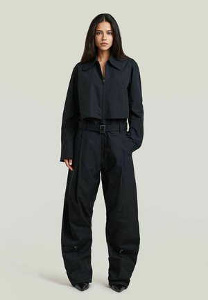 Femme aux longs cheveux foncés portant une veste courte noire et un pantalon large assorti avec une ceinture, debout devant un fond clair uni.