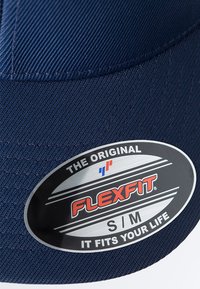 Navy cap with textured fabric, featuring a rounded crown and a flat brim. Includes a circular Flexfit label in black, red, and silver.