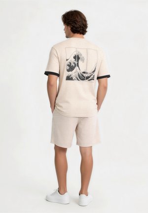 Muž stojí chrbtom, má na sebe béžové šortky a béžové tričko s čiernymi lemami rukávov a čiernym vlnitým vzorom na chrbte.