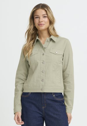 Jeune femme portant une veste boutonnée vert clair et un jean bleu foncé, debout devant un fond blanc uni.