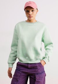 Sudadera verde menta con cuello redondo y puños acanalados. Combinada con pantalones cargo morados. Los accesorios incluyen una gorra rosa y aretes de aro.