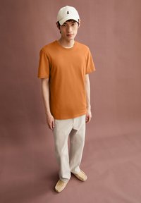Oranges Baumwoll-T-Shirt mit kurzen Ärmeln, kombiniert mit hellbeigen Hosen und beigen Slippern. Weiße Mütze mit schwarzem Logo.