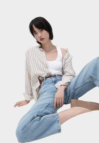 Junge Frau mit kurzem schwarzen Haar, die ein weißes Tanktop, ein gestreiftes beige Hemd, hochgeschnittene Jeans und braune Schuhe trägt, sitzt vor einem hellen Hintergrund.