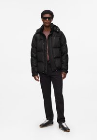 Sort puffjakke med hætte, med lynlås fortil og elastiske manchetter. Bæres over en pink top, sat sammen med sorte bukser og sorte sneakers.