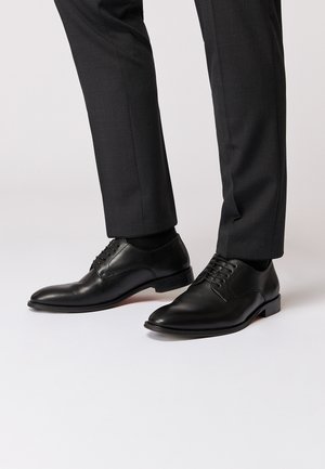 Schwarze Leder Schuhe mit einer glatten Oberfläche, spitzem Zeh und strukturierter Schnürung. Vor grauen Hosen und einem neutralen Hintergrund platziert.