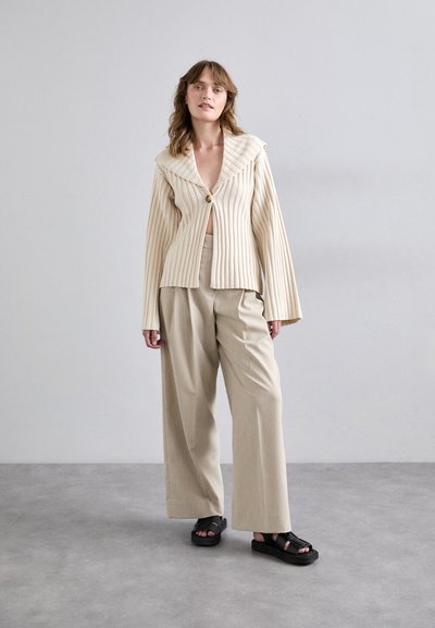 Beige ribbed blazer with large collar and single button, paired with loose beige trousers and black strap sandals on a gray background.