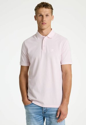 Junger Mann mit kurzen lockigen Haaren, der ein hellrosa kurzärmeliges Poloshirt und blaue Jeans trägt, vor einem schlichten weißen Hintergrund stehend.