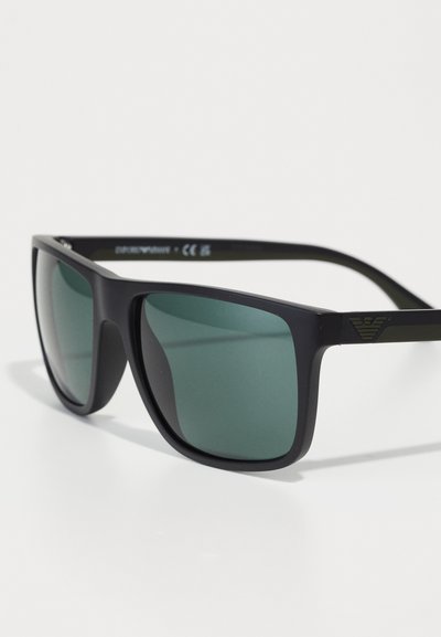 Ochelari de soare din plastic negru, cu lentile verde închis, formă pătrată, rame groase și detaliu subtil al logo-ului pe lateral.