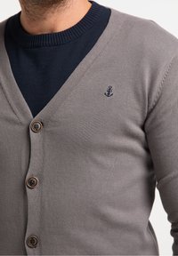 Cardigan grigio con scollo a V, dotato di quattro bottoni marroni e un piccolo emblema di ancora blu navy sul lato sinistro. Indossato sopra un maglione blu navy.