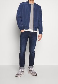 Chaqueta de vuelo navy con puños acanalados sobre un suéter gris a rayas. Jeans azul oscuro con los puños remangados y zapatillas deportivas grises.