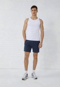 Débardeur en coton blanc, shorts navy, chaussures de sport avec accents argentés, debout contre un mur en briques blanches. Cheveux courts, pose neutre.
