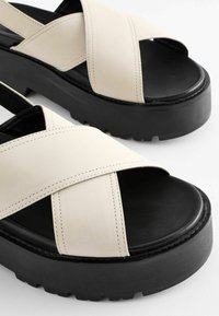 Beige sandals with cross straps and a chunky black platform sole with textured tread. The upper is made of smooth leather with contrasting stitching details.