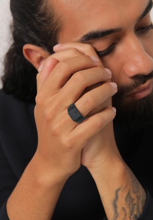 Must matte rõngas ruudukujuline. Sile tekstuur, mis kontrasteerub käte loodusliku nahatooniga. Peen pinnamuster on nähtav.