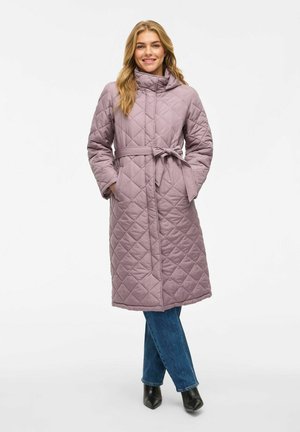 Žena oblečená v dlouhém prošívaném mauve kabátu s páskem, modrých džínách a černých botách na vysokém podpatku, stojící s rukama v kapsách.