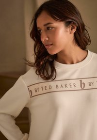 Baltas džemperis su grioveliais, ant krūtinės turintis horizontalią juostą su "b by TED BAKER" rudos spalvos rašmenimis.
