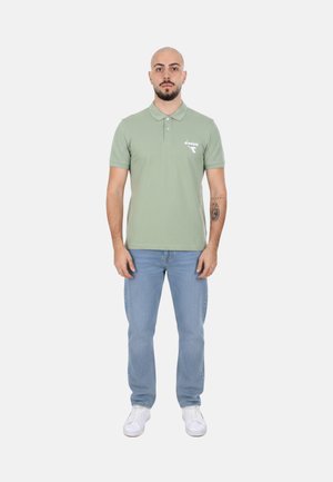 Uomo con barba e tatuaggio che indossa una polo verde chiaro, jeans azzurri e scarpe da ginnastica bianche, in piedi davanti a uno sfondo semplice.