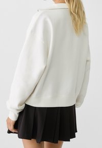 Sweat-shirt blanc avec col zippé et poignets côtelés, associé à une jupe plissée noire. Coupe ample, texture douce et design minimaliste.