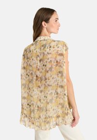 Mujer vistiendo una blusa sin mangas, de corte holgado, con un patrón abstracto multicolor y un dobladillo con volantes, mostrada desde atrás y en perfil lateral.