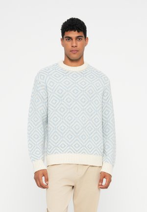 Young man wearing a light blue and white diamond-patterned knitted sweater and beige pants, standing against a white background.