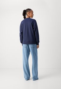 Ναυτικό φούτερ με μακριά μανίκια, στρογγυλή λαιμόκοψη και ριμπ πλέξη στους καρπούς, συνδυασμένο με ανοιχτό μπλε παντελόνι καμπάνα, παρουσιασμένο από πίσω σε ουδέτερο φόντο.