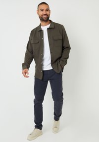 Chaqueta de camisa verde oliva con botones y dos bolsillos en el pecho, usada sobre una camiseta blanca y combinada con pantalones azul marino y zapatillas claras.