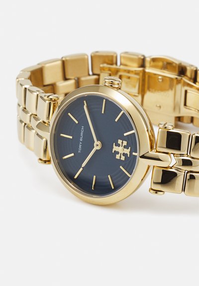 Orologio da polso in metallo dorato con quadrante nero, indici delle ore dorati, logo Tory Burch e cinturino a maglie su sfondo bianco.