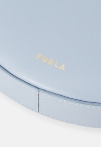 Svijetloplava kožna okrugla torba s zlatnim "FURLA" logom. Glatka tekstura s vidljivim šavom uz rub. Bez dodatnih hardverskih detalja.