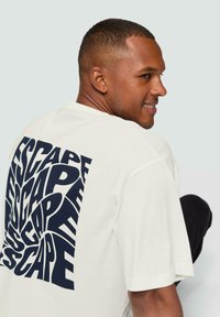 Witte katoenen t-shirt met een groot, vet navy blue grafisch patroon waarin het woord "ESCAPE" herhaald wordt, ronde halslijn, korte mouwen.