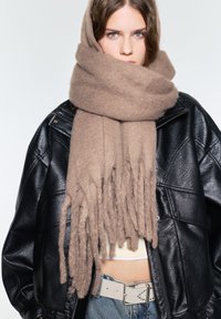 Beige wollen sjaal met rafelige randen, om de nek en het hoofd van het model gewikkeld, gelaagd over een zwart leren jasje en een lichte top.