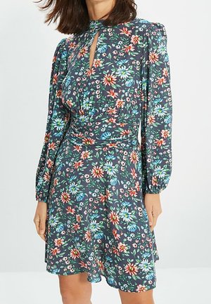 Kobieta ubrana w ciemną sukienkę z długim rękawem, sięgającą do kolan, z wielokolorowym kwiatowym wzorem i małym wycięciem w kształcie klucza z przodu.