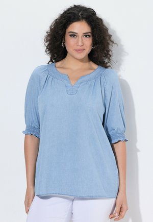 Kvinde med krøllet mørkt hår iført lyseblå bluse med smockede albuelange ærmer og hvide bukser, står mod en hvid baggrund.