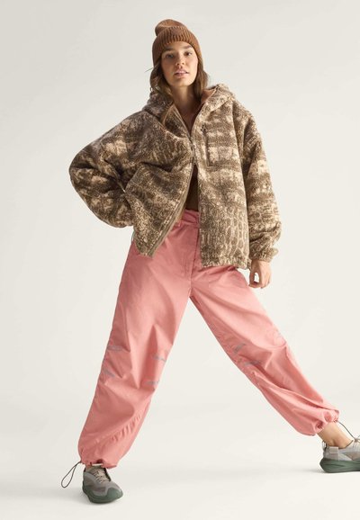 Fuzzy bruine zip-up jack met een patronenontwerp, gecombineerd met lichtroze tapered broeken met reflecterende accenten en grijze sportschoenen.