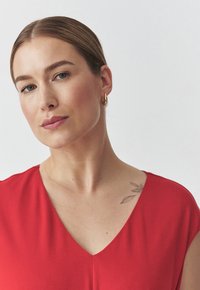 Punainen, hihaton mekko, jossa on syvä V-pääntie. Sujuva kangas, jossa on pehmeä rakenne. Olkapäällä on hienovarainen kukkatatuointi.