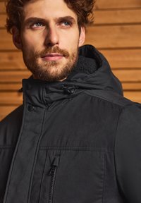 Černá zateplená bunda s vysokým límcem, zipovou přední kapsou, nastavitelnou kapucí a hladkým povrchem na dřevěném pozadí.